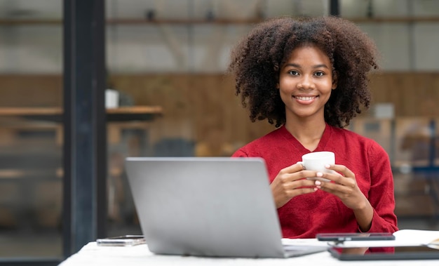 Portret van een lachende zwarte vrouw die aan een bureau zit en een koffiekopje vasthoudt en een laptop gebruikt die naar de gratis kopieerruimte van de camera kijkt