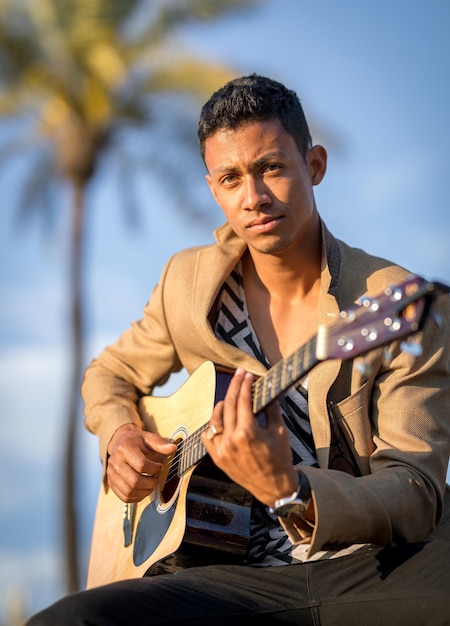 Portret van een Latijnse man die naar de camera kijkt en gitaar speelt op straat met palmbomen op de achtergrond bij zonsondergang