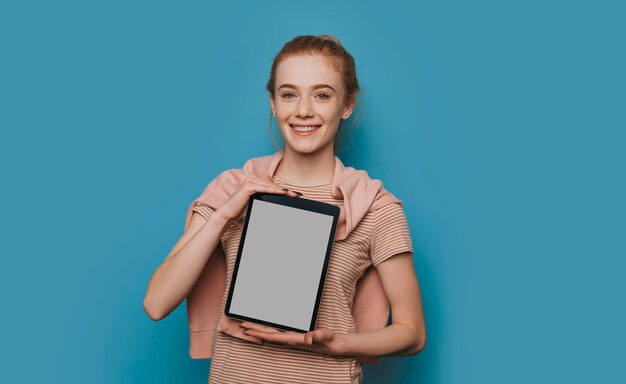 Portret van een leuke jonge vrouw met rood haar en sproeten die een tablet houden die een wit scherm toont die camera glimlachen bekijkt die op blauwe achtergrond wordt geïsoleerd.