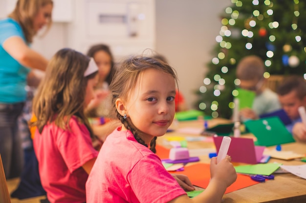 Portret van een lief klein meisje dat aan tafel zit met veel kleurrijke papieren terwijl ze naar de camera kijken Kinderen maken papieren kaarten tijdens de kerstworkshop