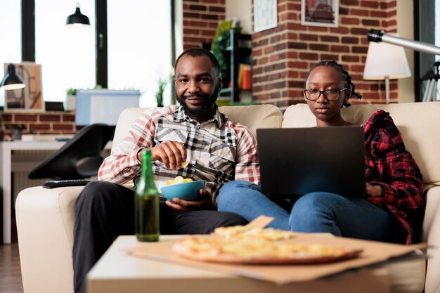 Portret van een man die tv-film kijkt en een vrouw die op laptop werkt, op internet surft en plezier heeft met film op televisie. Fastfoodmaaltijd eten van afhaalmaaltijden en flesjes bier drinken.