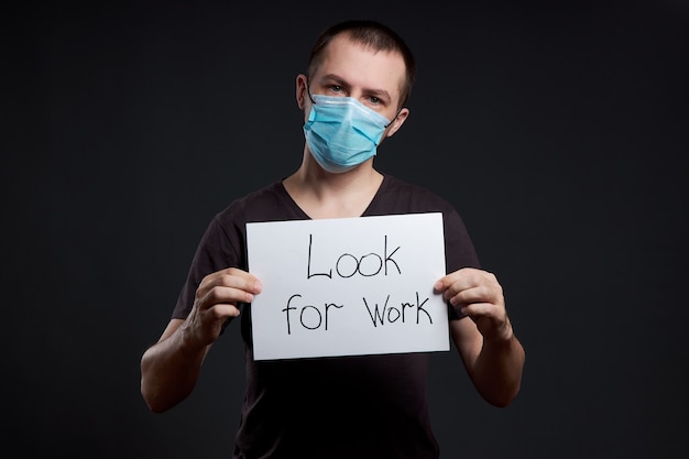 Portret van een man in een medisch masker met een bord op zoek naar werk op een donkere achtergrond, coronavirus-infectie