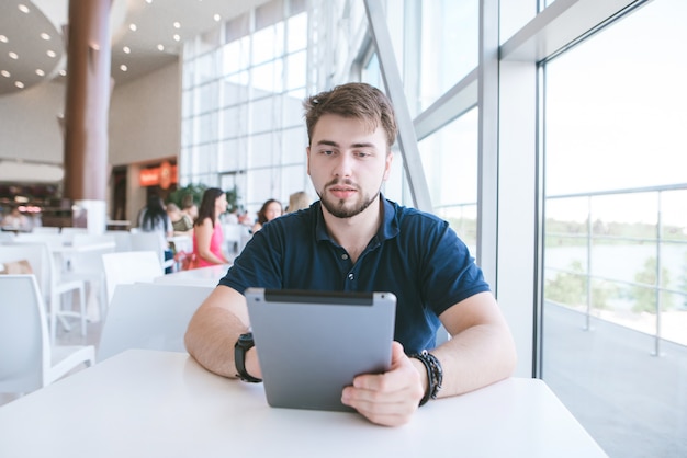 Portret van een man met een baard in casual kleding, zittend in een licht café en met behulp van een tablet.