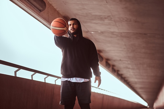 Portret van een man met een donkere huidskleur, gekleed in een zwarte hoodie en sportbroek, houdt een basketbal vast terwijl hij op een voetpad onder een brug staat.