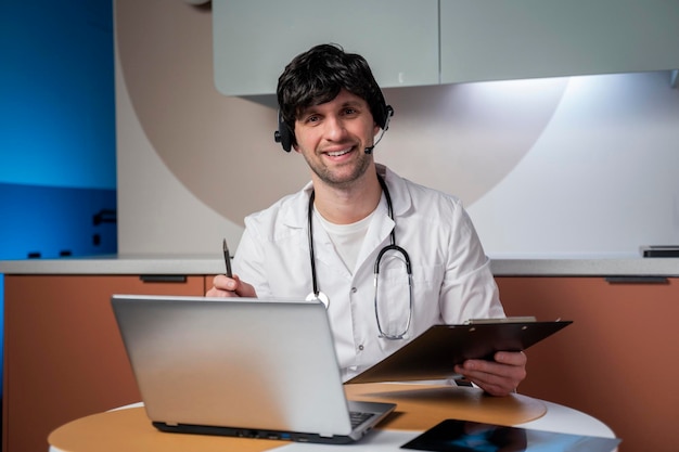 Portret van een mannelijke arts in zijn kantoor met behulp van een laptop om te videochatten met een patiënt online consultat