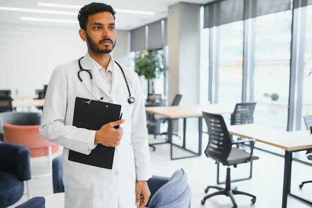 Portret van een mannelijke Indiase arts die een witte jas draagt met een open deur op de gang van de kliniek als achtergrond