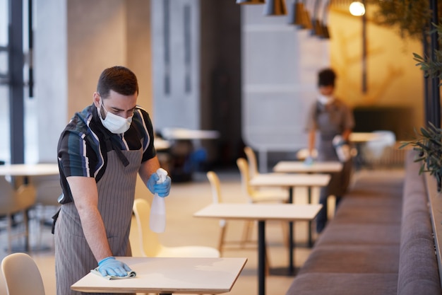 Portret van een mannelijke werknemer die een masker draagt tijdens het schoonmaken van tafels in het café-interieur, covid-veiligheidsconcept, kopieerruimte