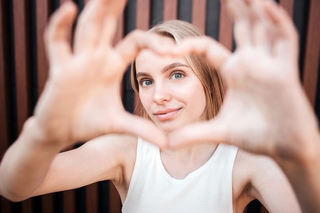 Portret van een meisje dat een hart van haar vingers maakt en het toont. Ook kijkt ze ernaar en glimlacht.