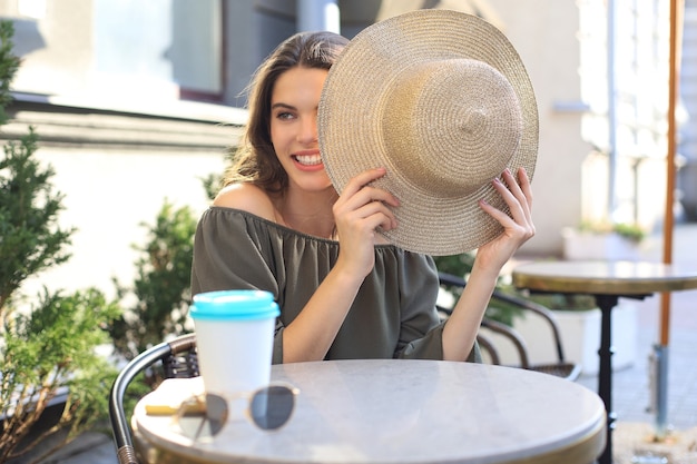 Portret van een meisje dat haar gezicht achter een strohoed verbergt terwijl ze in het zomerstraatcafé zit.