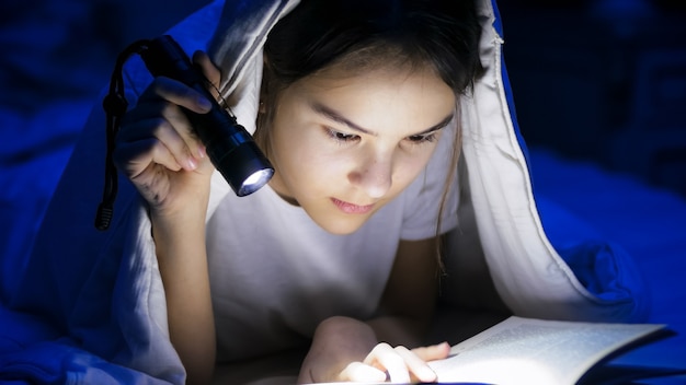 Portret van een meisje dat 's nachts onder de deken ligt en een boek leest met een lichte fakkel.