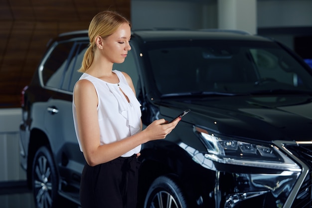 Portret van een meisje in een autodealer met een telefoon in haar handen op de achtergrond van een auto