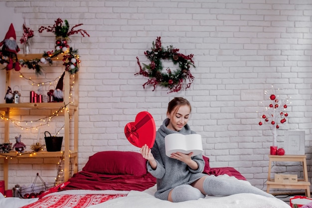 Portret van een meisje in een grijze sweater met een hart-vormige rode giftvakje zitting in de flat, de dagconcept van Valentine, exemplaarruimte