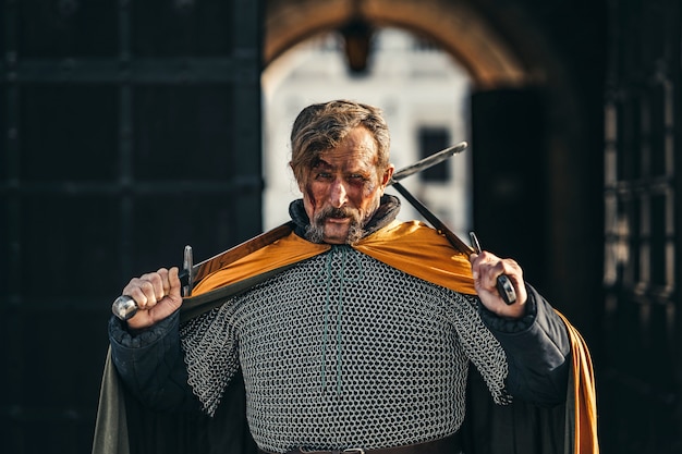 Portret van een middeleeuwse senior krijger in harnas na een gevecht met bloed op zijn gezicht. De krijger houdt twee zwaarden in zijn handen