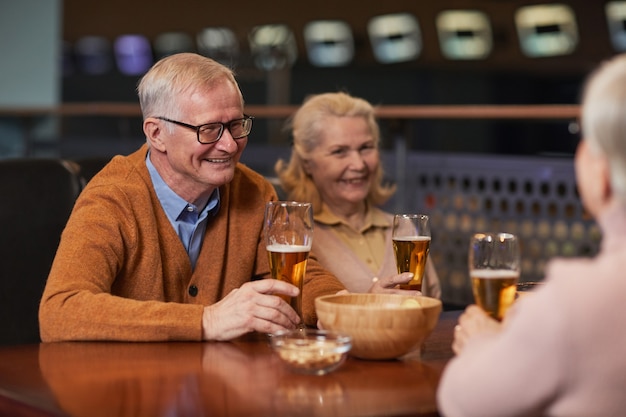 Portret van een modern senior koppel dat bier drinkt in de bar en lacht terwijl ze genieten van een avondje uit met vrienden, kopieer ruimte