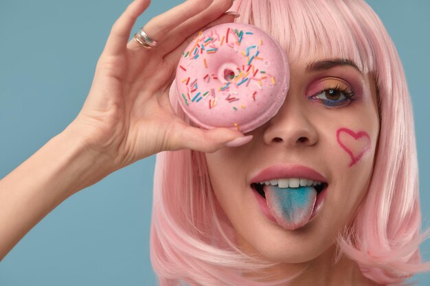 Portret van een modieus meisje met een roze pruik en met een roze donut in haar handen houdt de tong in de gaten