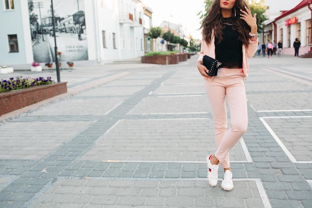 Portret van een modieus trendy meisje met kapsel, gekleed in een stijlvol sport-chic pak van roze kleur en zwarte kanten top die op straat staat met een handtas in de hand. kopieerruimte.