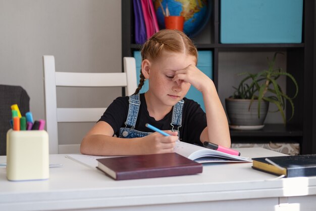 Portret van een moe, uitgeput Kaukasisch roodharig meisje dat verdrietig kijkt terwijl ze aan het bureau zit en thuis huiswerk maakt. Wanhoop. Online onderwijs. Quarantaine. Terug naar schoolconcept.