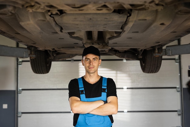 Portret van een monteur die repareert in uniform staande camera met gekruiste handen naar een onder opgeheven auto