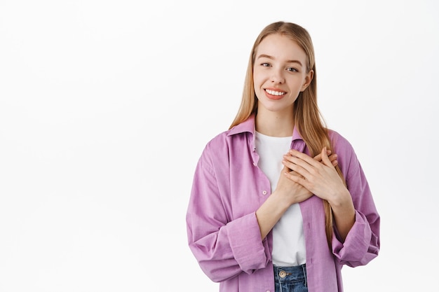 Portret van een mooi blond meisje dat zich aangeraakt en dankbaar voelt, kijkend met een oprechte glimlach, hand in hand op het hart, hartverwarmend moment, staande over een witte muur