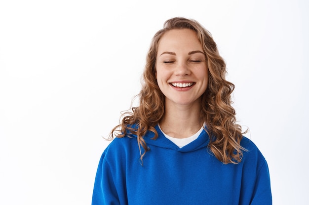 Portret van een mooi blond vrouwelijk model sluit de ogen, glimlacht tevreden, herinnert zich een gelukkig moment, dagdromen, staande tegen een witte muur