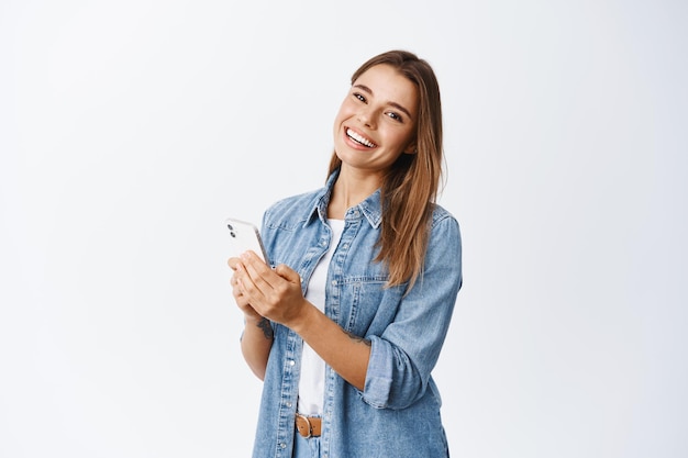 Portret van een mooi duizendjarig meisje met een witte glimlach, berichten verzenden, chat-app gebruiken op smartphone en kijken, staande over wit