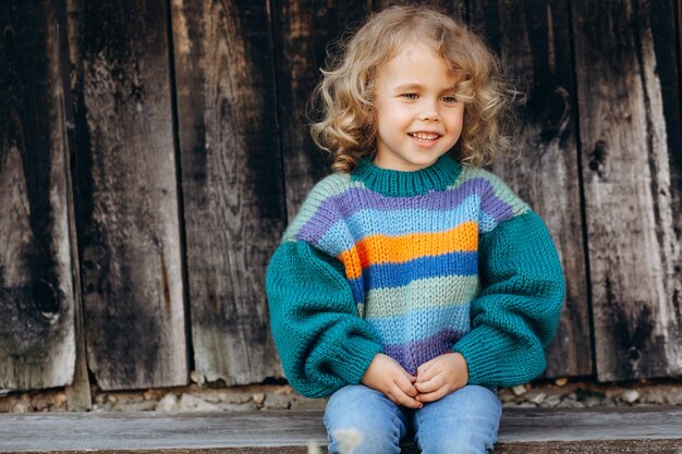 Portret van een mooi en gelukkig krullend meisje in een gebreide trui in de buurt van een houten muur