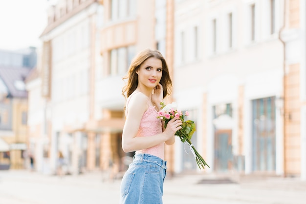 Portret van een mooi en mooi meisje in de straat van de stad, badend in de ondergaande zon. Meisje dat een boeket rozen houdt