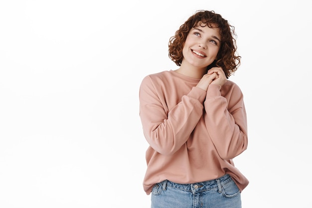 Portret van een mooi jong meisje dat aan het dagdromen is, naar de top kijkt met liefde en romantische emotie, handen bij elkaar houdt en omhoog staart, gebiologeerd door iets moois, witte achtergrond.