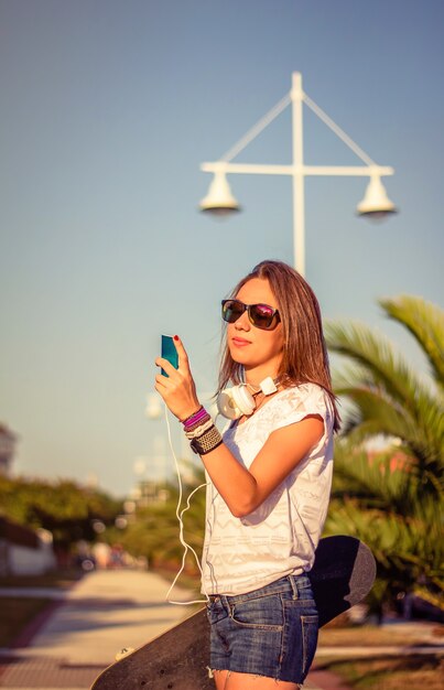 Portret van een mooi jong meisje met skateboard en koptelefoon op zoek naar haar smartphone buitenshuis. Warme tinten editie.