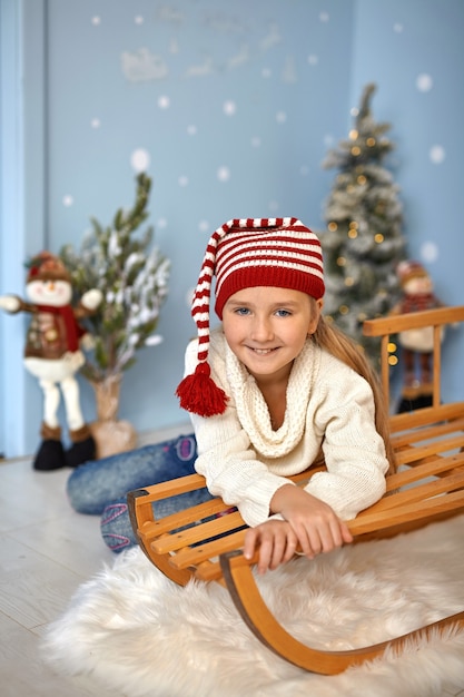 Portret van een mooi kindmeisje met een rode hoed in een kerstinterieur zittend op een houten slee omringd door speelgoed. Fijne vakantie, blij kind. Klein kind viert Kerstmis en Nieuwjaar.