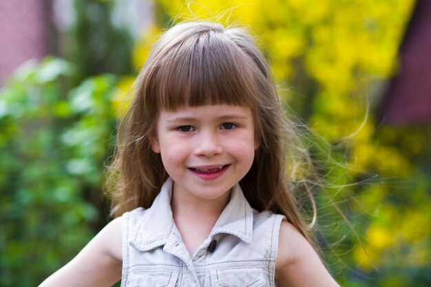 Portret van een mooi klein langharig blond peutermeisje in mouwloze witte kleding die verlegen in camera tegen vaag in openlucht glimlachen. Onschuldig gelukkig jeugdconcept.