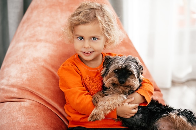 Portret van een mooi klein meisje dat op een oranje stoel zit met een hond in haar armen die naar de camera kijkt en glimlacht