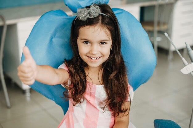 Portret van een mooi klein meisje duim opdagen na een tandoperatie. Gelukkig kind na tandonderzoek in een pediatrische stomatologie.