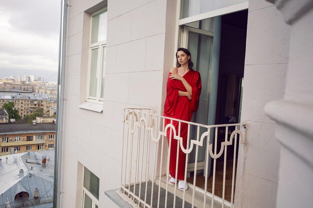 Foto portret van een mooi meisje dat in de zomer op het balkon van een hoog gebouw in een rode badjas staat