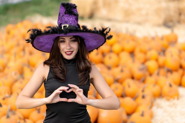 Portret van een mooi meisje in een zwarte jurk en een heksenhoed Poseren tegen de achtergrond van pompoenen Toont een hart met zijn handen Halloween-pompoendecor