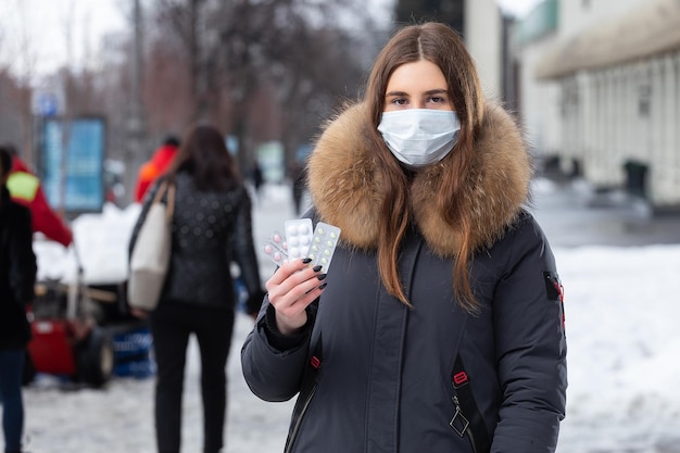 Portret van een mooi meisje met een medisch beschermend masker in het houden van witte pillen in haar handen voor verkoudheid en griep Winter straatportret van een vrouw onder sneeuwval