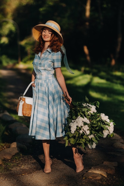 Portret van een mooi meisje met lang haar, een strohoed en een lange zomerjurk met lila bloemen in de tuin