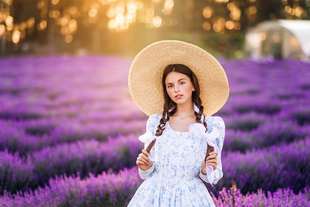 Portret van een mooi meisje op een lavendelachtergrond. Ze draagt een witte jurk en een grote hoed. Zomerfoto in de zon