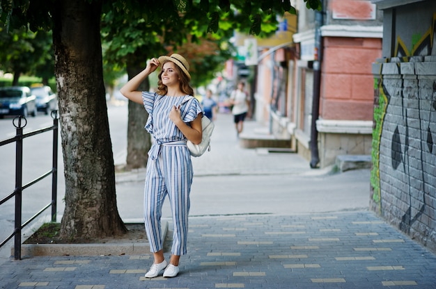 Portret van een mooi model in gestreepte algehele poseren met hoed en een rugzak op een straat met bomen in een stad.