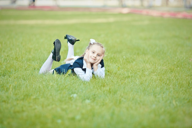 Portret van een mooi schoolmeisje met een vlecht en een witte boog die op het groene gras in het park ligt