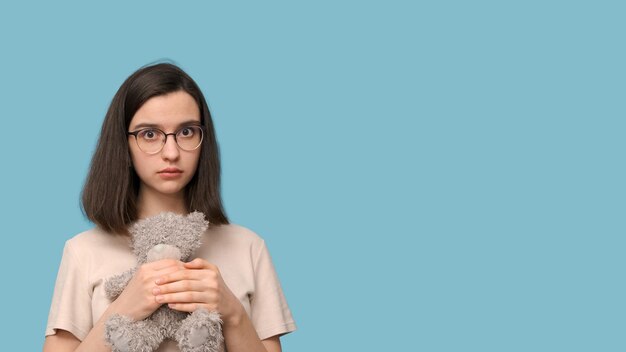 Portret van een mooi studentenmeisje met een bril die een grijs teddybeerspeeltje knuffelt op een blauwe achtergrond