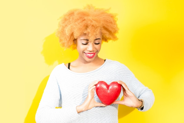 Portret van een mooie Afrikaanse vrouw die rood hart op de gele achtergrond houdt. Valentijnsdagconcept
