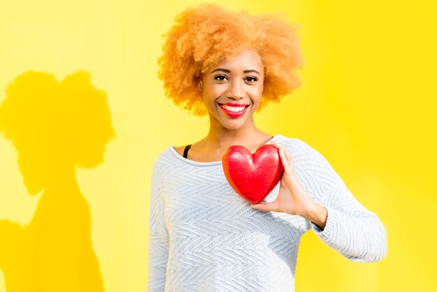 Portret van een mooie Afrikaanse vrouw die rood hart op de gele achtergrond houdt. Valentijnsdagconcept