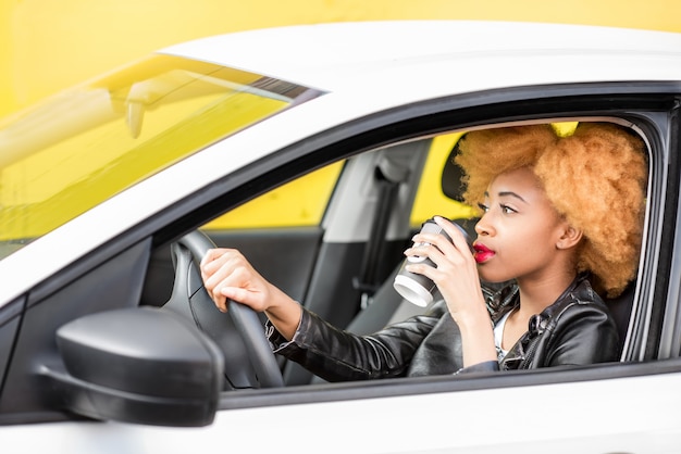 Portret van een mooie Afrikaanse vrouw in leren jas met koffiekopje zittend in de auto op de gele achtergrond