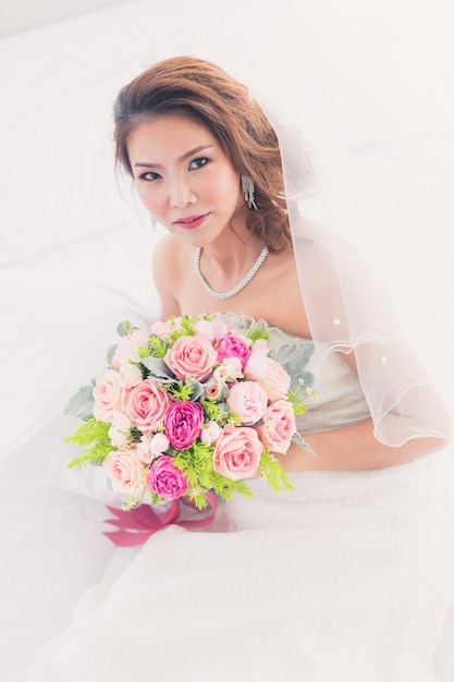 Portret van een mooie Aziatische bruid die in de kleedkamer zit en een boeket bloemen vasthoudt met een witte achtergrond.