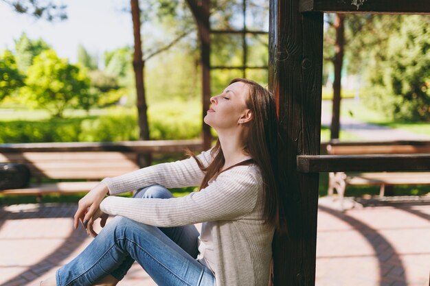 Portret van een mooie blanke jonge vrouw met een charmante glimlach die lichte vrijetijdskleding draagt. Lachende vrouw zittend onder boog in stadspark in straat buiten op lente natuur. Lifestyle-concept