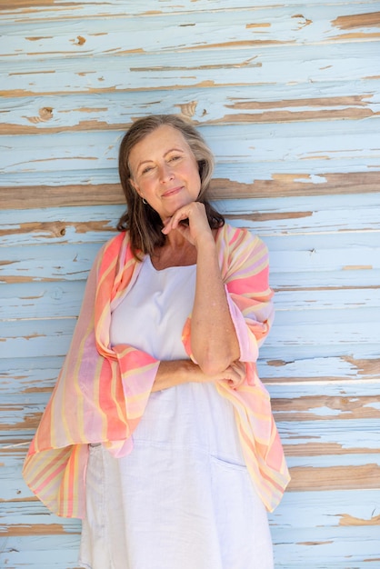 Portret van een mooie blanke senior vrouw met de hand op de kin die tegen een houten muur staat. Kopieer ruimte, ongewijzigd, pensioen, vakantie, sjaal, levensstijl, glimlachen en plezierconcept.