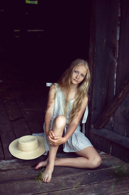 Portret van een mooie blonde dorpsmeisje met lang haar in een jurk en hoed zittend bij een houten oude schuur in de zomer