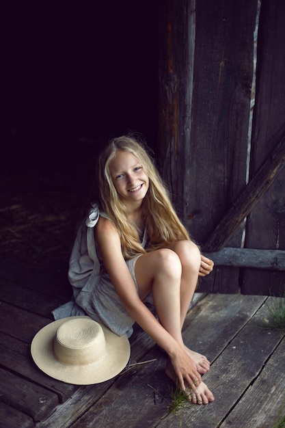 Portret van een mooie blonde dorpsmeisje met lang haar in een jurk en hoed zittend bij een houten oude schuur in de zomer