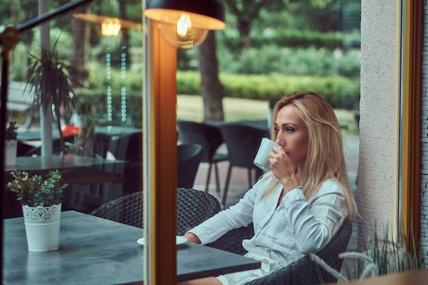 Portret van een mooie blonde vrouw gekleed in een witte blouse zittend aan een tafel drinkt koffie op terras café.